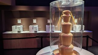 Five chocolate tasting machines, each with a description of the chocolate they contain hanging on the wall above them. In front of the machines is a large chocolate fountain with caramel coloured chocolate.