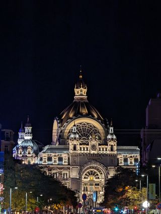 The Antwerp Central train station building at night. The facade of the train station is lit up with white lights that reflect off the gold fixtures on the extorior of the building. The dome at the top of the station is illuminated with a soft yellow light.