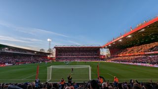 Beyond the goal which is situated in front of the away fans, the players of Brighton and Nottingham Forest prepare to kick-off. The home fans in the three other stands that are visible, are holding red and white scarves above their heads.