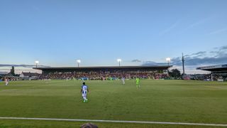 View of the pitch from the terrace in the away end at The Bolt New Lawn Stadium. Brighton are playing in blue and white striped shirts with white shorts and Forest Green Rovers are wearing lime green shirts that have black 'tiger' stripes and lime green shorts.