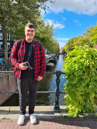 Declan wearing a red and black checked shirt over a black t-shirt with black jeans and white trainers, holding his camera, and stood in front of some railings on a bridge over the canal. To the right of Declan is a plant with lime green leaves which blend into the leaves of the trees which line either side of the canal in the background.