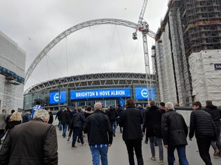 Group of people walking towards Wembley Stadium (London) with a Brighton and Hove Albion banner shown on the outside of the stadium.