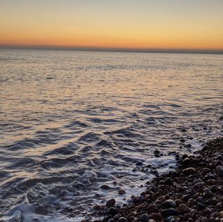 Calm sea with small wave breaking onto shingle and an orange sky.