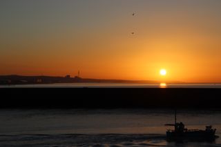 Sun rising above a harbour wall reflecting on the water. A fishing boat is in front of the harbour wall. East Sussex coastline in the distance.
