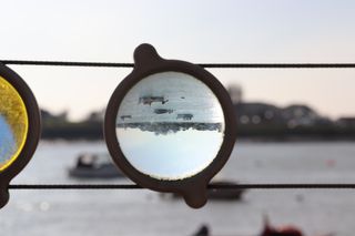 A circular glass panel attached to some railings. Within the glass panel, an upside down view of small boats on a river.