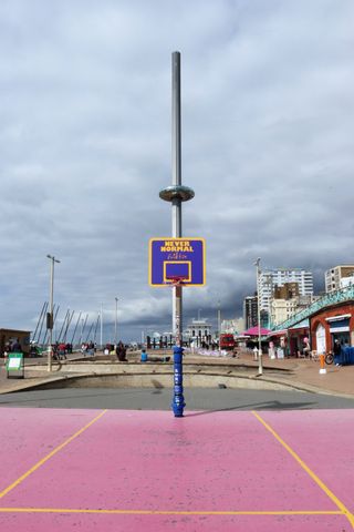 Basketball hoop on the western side of the basketball court on Brighton beach. Behind and aligned with the hoop, the I360 is ascending.