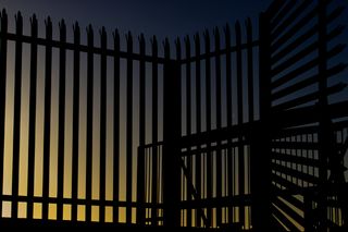 Spiked railings and a gate that overlap with a blue/yellow gradient sky behind the railings.