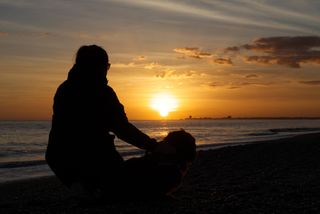 A woman with her hair in a ponytail kneeling next to a Sussex spaniel on a pebble beach while watching the sunset.