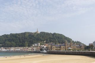 The eastern side of La Concha beach in San Sebastián. The tide is out which exposes the gold coloured sand. Above the sea wall which lines the beach is San Sebastián city hall. Behind the city hall is the old town of San Sebastián which is overlooked by the tree covered hill of Monte Urgull. At the top of Monte Urgull is a statue of Jesus.